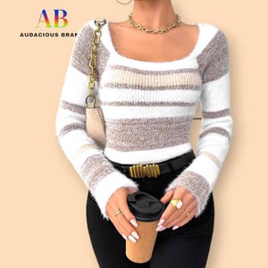May include: A fluffy, striped sweater with a square neckline. The sweater has horizontal stripes in white, beige, and brown. The image also shows a black belt with a gold buckle, a gold necklace, and a coffee cup.