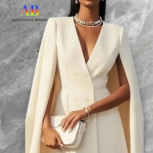 Puede incluir: Vestido blazer de doble botonadura color marfil con mangas capa. El vestido presenta un escote en V y está adornado con un collar y una pulsera dorados. Se sostiene un bolso blanco. La marca "AUDACIOUS BRAND" es visible.