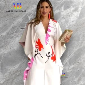 Puede incluir: Un vestido blanco estilo caftán con cuello en V con un diseño floral en rojo y rosa. El vestido tiene mangas anchas y un ribete rosa. La imagen incluye el texto "AB Audacious Brand".