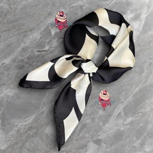 May include: A black and white patterned silk scarf with a geometric design. The scarf is tied in a knot and the ends are hanging down.