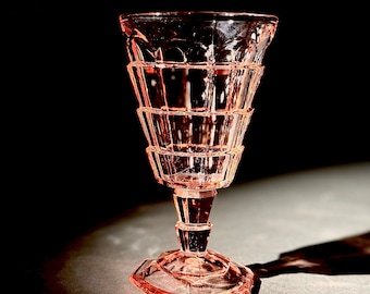 May include: A pink-tinted, vintage-style glass goblet with a faceted design. The goblet features a flared bowl, a stem with a decorative element, and a hexagonal base. The glass is transparent, allowing light to pass through, and is set against a dark background.