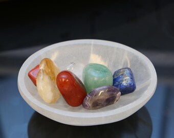 May include: A white selenite bowl filled with seven chakra stones. The stones are red, orange, yellow, green, blue, indigo, and violet.