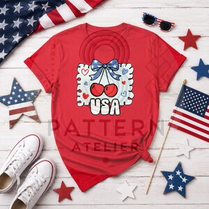 Puede incluir: Una camiseta roja con un gráfico de cerezas, un lazo azul y la palabra "USA". El diseño está rodeado de corazones y elementos decorativos. La imagen también incluye una bandera estadounidense, gafas de sol y decoraciones en forma de estrella.