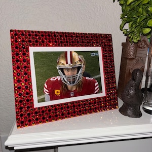 May include: A red picture frame with a sparkling rhinestone border. The frame holds a photo of a football player in a red and gold uniform. The photo is set against a white background.