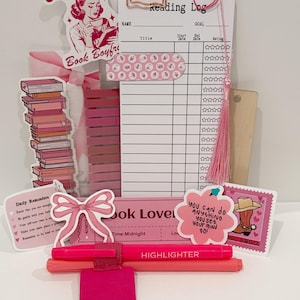 May include: A reading log planner set featuring a white card with a grid, a pink tassel, and various stickers including a stack of books, a crying person with the text Crying Over Book Boyfriends, a pink bow, and a stamp with a cowboy boot.
