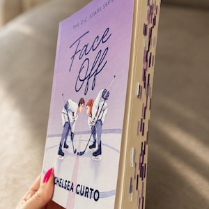 May include: A paperback book titled Face Off by Chelsea Curto from The D.C. Stars Series. The cover features an illustration of two hockey players on ice. The book edges are decorated with purple and white page markers in a vertical pattern.