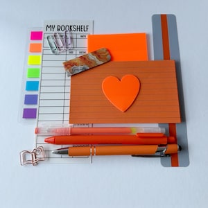 May include: A collection of stationery including a My Bookshelf tracker, neon sticky notes, a marbled rectangular piece, a lined orange card with a heart cutout, three pens in shades of orange, and rose gold paper clips on a gray ruler.