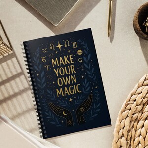 May include: A spiral-bound notebook with a dark blue cover featuring the text "MAKE YOUR OWN MAGIC" in gold lettering. The design includes celestial symbols, hands, and botanical elements. The notebook is open, revealing the spiral binding.