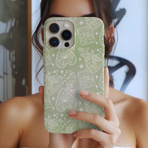 Puede incluir: Una funda de teléfono verde salvia con un patrón blanco repetido de hojas de cannabis, polillas, estrellas y frascos. La funda tiene un acabado brillante y un recorte para la cámara.