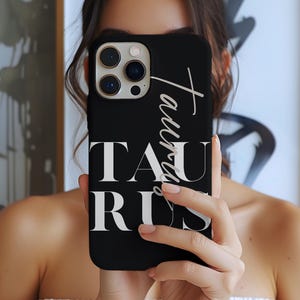 May include: Black phone case with the word "Taurus" in white script and block letters. The phone case has a camera module with three lenses and a flash. The case is held in front of a person.