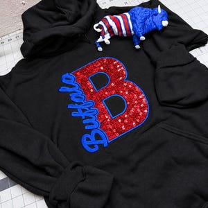 May include: A black hooded sweatshirt featuring a large red sequined letter B with a blue embroidered outline and the word Buffalo in blue script. A small blue, white, and red buffalo figurine rests on the shoulder area of the hoodie.