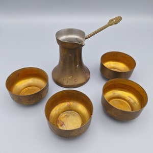Puede incluir: Un juego de cuatro pequeñas tazas de latón y una cafetera de latón con asa. Las tazas y la cafetera tienen un aspecto desgastado y antiguo.