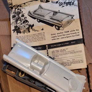 Puede incluir: Un kit de modelo de coche vintage con carrocería de plástico blanco, parcialmente ensamblado, junto a un anuncio retro. El anuncio presenta un dibujo del coche y el texto "TIGER SPORT CAR". El kit incluye varias piezas de plástico gris.