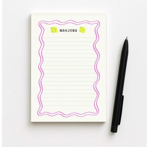 May include: A lined notepad with a pink wavy border and the word "MAHJONG" at the top. The notepad has a white background and a black pen is placed to the right of the notepad.