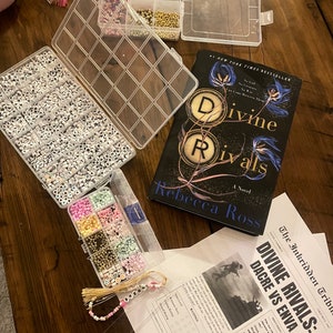 May include: A close-up of a book titled "Divine Rivals" by Rebecca Ross, a clear plastic container filled with white beads with black letters, a clear plastic container filled with colorful beads, and a beaded bookmark with the name "BREN" on it.