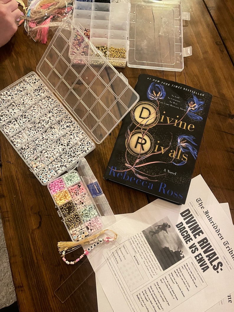 May include: A close-up of a book titled "Divine Rivals" by Rebecca Ross, a clear plastic container filled with white beads with black letters, a clear plastic container filled with colorful beads, and a beaded bookmark with the name "BREN" on it.