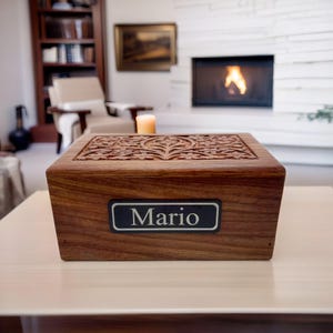 May include: A wooden cremation urn with an intricate carved design on the lid. The urn is a dark reddish-brown color and has a silver nameplate that reads "Mario".