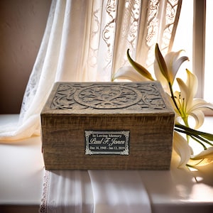 May include: A wooden cremation urn with a carved floral design. The urn has a black label with silver lettering that reads "In Loving Memory Paul F. Jones Dec 14, 1945 - Jan 12, 2019".