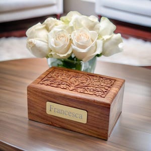 May include: A wooden cremation urn with an intricate carved design and a brass nameplate that reads "Francesca". White roses are in the background.