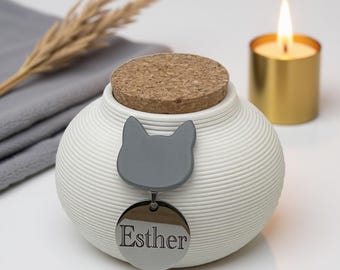May include: A white, ribbed ceramic pet urn with a cork lid. A gray cat-shaped emblem and a silver tag with the name "Esther" are attached. A lit candle in a gold holder is in the background.