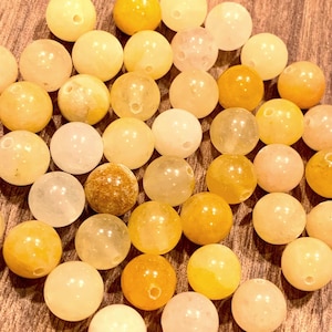 May include: A collection of round beads in various shades of yellow, cream, and amber. The beads have a smooth, polished surface and a small hole drilled through the center. The beads are scattered on a wooden surface.