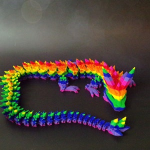 May include: A rainbow-colored dragon figurine made of plastic. The dragon is curled up in a circle with its tail in its mouth. The dragon is made up of many small, triangular pieces that are arranged in a pattern to create the rainbow effect.