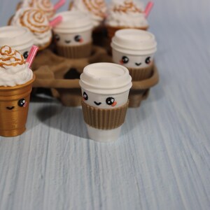 May include: A collection of miniature coffee cups with cute faces. Some cups have whipped cream and caramel drizzle, others have lids. All have brown sleeves and pink straws. The cups are arranged in a brown cardboard carrier.