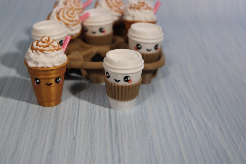May include: A collection of miniature coffee cups with cute faces. Some cups have whipped cream and caramel drizzle, others have lids. All have brown sleeves and pink straws. The cups are arranged in a brown cardboard carrier.