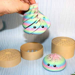 May include: A rainbow-colored, articulated dumpling toy with a cute face featuring black eyes and pink cheeks. The toy is shown in two parts, with one piece held up and another resting on a surface, accompanied by two tan, round steamer baskets.