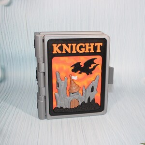 May include: A gray, hinged toy with a black and orange front panel. The panel features the word "KNIGHT" in orange letters, a black dragon, and a castle illustration. The toy is designed to open and close.