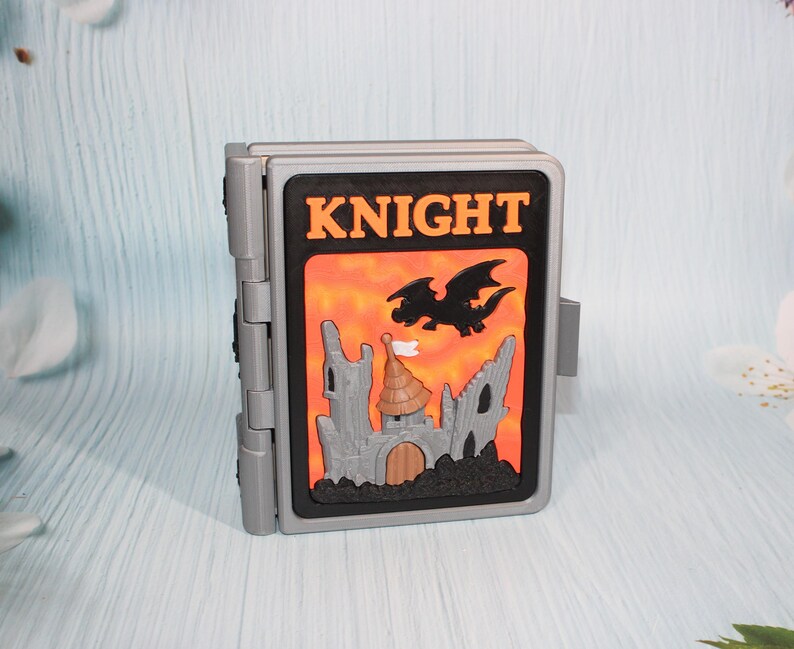 May include: A gray, hinged toy with a black and orange front panel. The panel features the word "KNIGHT" in orange letters, a black dragon, and a castle illustration. The toy is designed to open and close.
