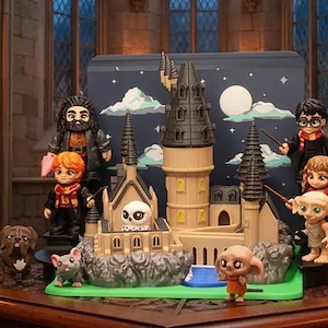 May include: A miniature plastic castle playset featuring a central tower, stone-like base, and multiple small character figurines including a bearded man, a boy with glasses, a girl, two house-elves, a dog, and a small owl, set against a night sky backdrop.
