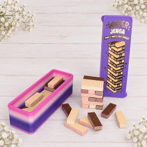 May include: A purple and pink gradient rectangular container and a matching purple box labeled Wafer Jenga, Stack It, Snack It, Don't Crumble! with a graphic of stacked wafers. Several rectangular wafer cookies are scattered on the surface.
