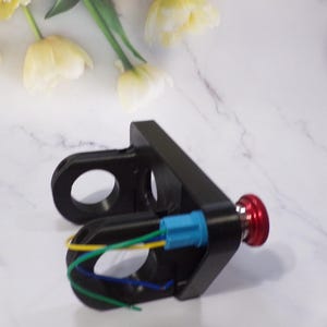 May include: Black metal component with two circular openings and a red button. It has a blue connector and colored wires. The component is on a white surface with yellow and white flowers in the background.