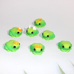 May include: A collection of eight small, green frog figurines with black eyes. Some frogs have decorative accents like flowers, horns, or bows. The frogs are arranged on a white surface.
