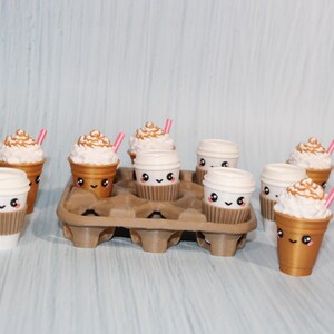 May include: A collection of miniature coffee cups, some with whipped cream and pink straws, arranged in a brown cardboard carrier. The cups have cute, smiling faces. The background is a light blue.