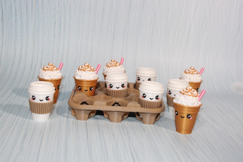 May include: A collection of miniature coffee cups, some with whipped cream and pink straws, arranged in a brown cardboard carrier. The cups have cute, smiling faces. The background is a light blue.