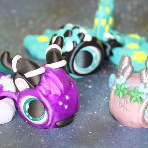May include: Three small, colorful, hand-painted figurines of mythical creatures. The first is a purple dragon with black and white accents, the second is a teal and black dragon with a turquoise eye, and the third is a pink and silver creature with green accents and a green eye.