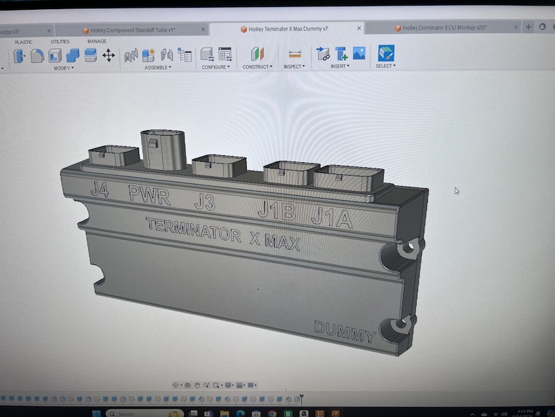 May include: A gray 3D model of an electronic control unit (ECU) with the text "TERMINATOR X MAX" and "DUMMY" printed on it. The ECU has multiple connectors labeled "J4", "PWR", "J3", "J1B", and "J1A".