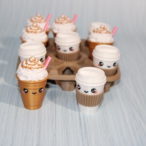 May include: A collection of miniature coffee cups and frappes with cute, smiling faces. The frappes have whipped cream and pink straws. The cups are in a brown cardboard carrier, set against a light blue background.