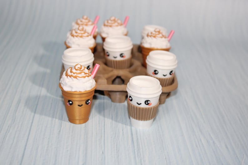 May include: A collection of miniature coffee cups and frappes with cute, smiling faces. The frappes have whipped cream and pink straws. The cups are in a brown cardboard carrier, set against a light blue background.