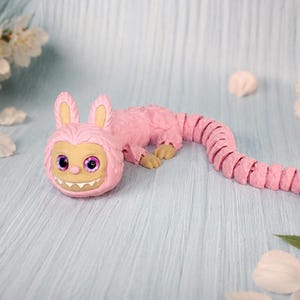 May include: A pink articulated creature figurine with long rabbit-like ears, large purple eyes, and a wide grin showing white teeth. The body features a textured, segmented design that extends into a long, flexible tail with tan-colored feet.