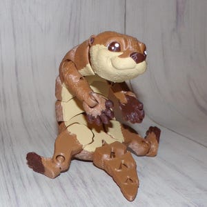 May include: A brown and tan articulated toy otter with a smiling expression. The toy has a segmented body and limbs, with dark brown accents on the paws and nose. The otter is posed sitting upright.