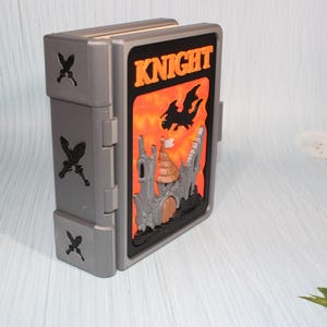May include: A gray, hinged box with a fantasy-themed design. The front features a castle, dragon, and the word "KNIGHT" against an orange and black background. Decorative crossed swords are on the side.