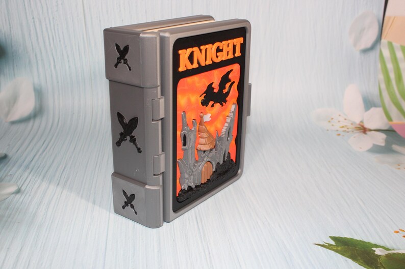 May include: A gray, hinged box with a fantasy-themed design. The front features a castle, dragon, and the word "KNIGHT" against an orange and black background. Decorative crossed swords are on the side.