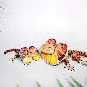 May include: Two toy frilled-neck lizards are displayed on a white surface, with green leaves in the background. The lizards have tan heads, yellow and red frills, and striped bodies. The frills have a spotted pattern.