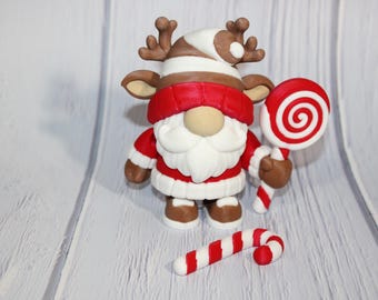 May include: A festive Christmas gnome figurine with reindeer antlers, a red and white outfit, and a long white beard. The gnome holds a red and white swirled lollipop and a candy cane. The figurine is a holiday decoration.
