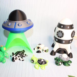 May include: A collection of whimsical space-themed figurines. Includes a gray and blue UFO with a green light, a white and black rocket, and several turtle-shaped aliens in various colors and patterns. The figurines are displayed on a light blue surface.