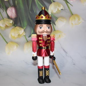 May include: A decorative nutcracker figurine in a regal design. The figure is predominantly red with gold accents, a black belt, and white pants. It wears a crown and holds a golden sword. The nutcracker is a classic holiday decoration.