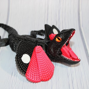 May include: A black and red dinosaur figurine with an open mouth, showing its red tongue and white teeth. The dinosaur has orange eyes and white spots on its head. The figurine is lying on a white wooden surface.